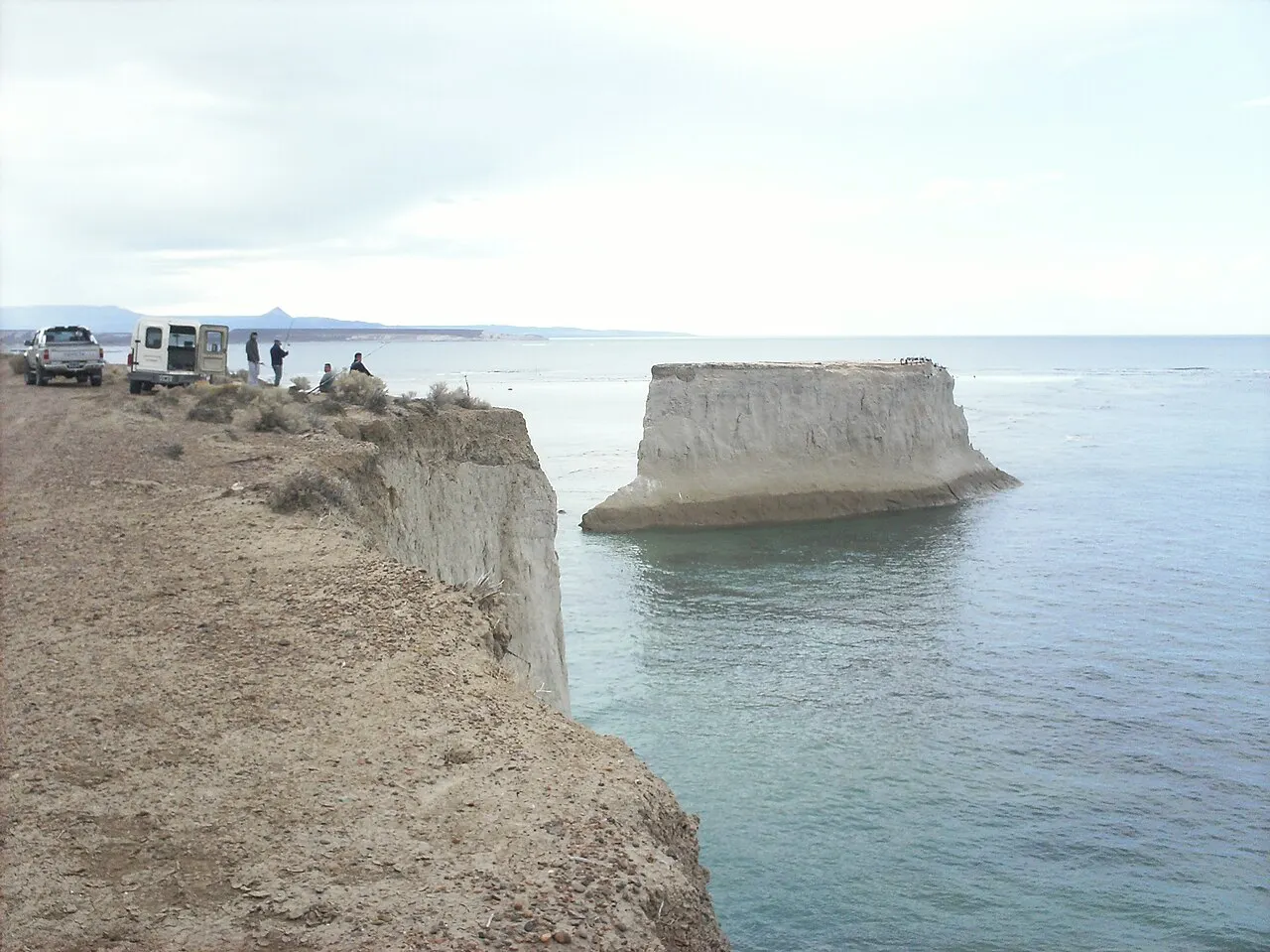 El farallón junto al Faro San Jorge: una columna de roca separada del acantilado, con aves marinas arriba, y pescadores en el borde de la meseta