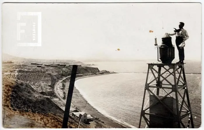 El primer faro de Comodoro, hacia 1920: una estructura de hierro con la linterna arriba y un farolero, con la bahía y la ciudad naciente detrás