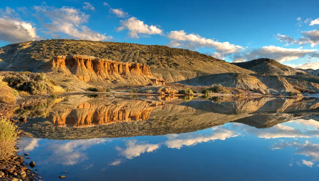 Barranca de roca roja tallada por la erosión en el Área Natural Protegida Rocas Coloradas, Chubut, reflejada en agua quieta al atardecer entre lomas de estepa patagónica